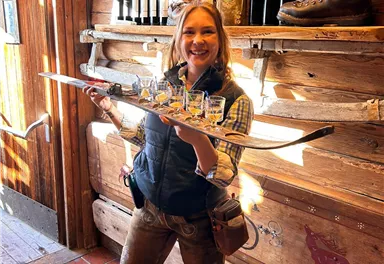 Eine fröhliche Frau in traditioneller bayerischer Tracht hält ein Skischuh mit Gläsern in der Hand. Im Hintergrund sind Weinflaschen und eine rustikale Holzverkleidung zu sehen.