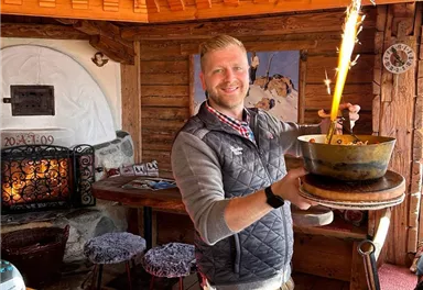 Ein fröhlicher Kellner trägt ein Gericht mit sprühenden Funken in einem holzgetäfelten Restaurant. Im Hintergrund sieht man eine gemütliche Atmosphäre mit einem Kamin und Holzstuhl.
