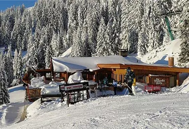 Eine gemütliche Berghütte im Schnee, umgeben von hohen, schneebedeckten Bäumen. Die Sonne scheint auf die Winterlandschaft und schafft eine einladende Atmosphäre.