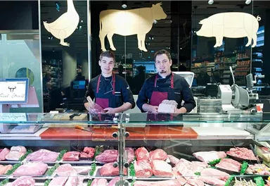 Zwei Metzger stehen hinter einer Theke mit frischem Fleisch. Die Auslage zeigt eine Vielzahl von Fleischsorten in einer modernen Metzgerei.