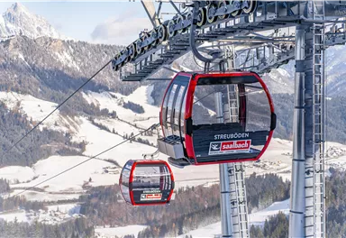 Zwei moderne Gondeln fahren auf einer Seilbahn über schneebedeckte Berge. Im Hintergrund sind majestätische Berglandschaften zu sehen.