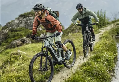 Two cyclists are riding on a narrow path in the mountains. The landscape is green and hilly, with an impressive mountain in the background.