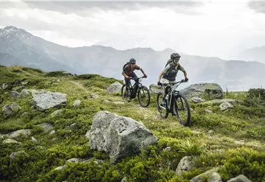 Two cyclists are riding on a forest path through a mountainous landscape. In the background, the mountains and a cloudy sky can be seen.