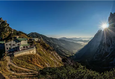 Eine beeindruckende Berglandschaft mit einem Gasthaus auf einer Terrasse. Die Sonne geht am Horizont auf und beleuchtet das Tal.