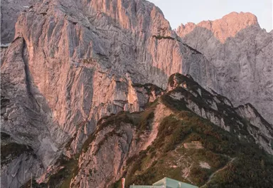 Eine beeindruckende Berglandschaft mit steilen Felsen und einem Hüttengebäude im Vordergrund. Die Felsen sind in sanften Abendlichttönen beleuchtet.