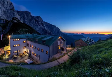 Ein malerisches Berghotel in der Dämmerung, umgeben von majestätischen Bergen. Die Fenster strahlen warmes Licht aus und schaffen eine einladende Atmosphäre.