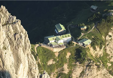 Eine abgelegene Hütte in den Bergen, umgeben von steilen Felsen und grünem Gelände. Die Dächer sind grün und die Landschaft ist reichhaltig und naturbelassen.