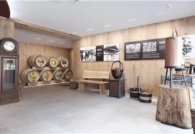 Ein Raum mit Holzwänden und Boden, in dem historische Fässer an einer Wand stehen. Es gibt Sitzgelegenheiten und Ausstellungsstücke zur Geschichte.