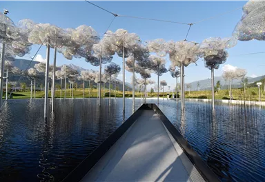 A landscape with a water basin and artistic, cloud-like installations. In the background, mountains and a clear sky are visible.