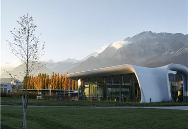 A modern building with a curved shape stands in a green park. In the background, majestic mountains and a clear sky can be seen.