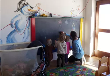 Ein helles Klassenzimmer mit einer Wandtafel, an der eine Person und zwei Kinder zeichnen. An der Wand ist ein fröhlicher Schneemann gemalt.