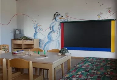 Ein fröhlicher Raum mit einem Tisch und Stühlen, ideal für Kinder. An der Wand sind lustige Schneemänner gemalt, und es gibt ein großes schwarzes Board.