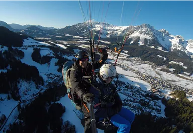 A tandem flight with paragliding over snow-covered mountains. In the background, picturesque valleys and a clear blue sky can be seen.