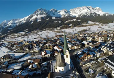 A picturesque mountain landscape with snow-covered peaks and a charming village in the foreground. The church with the tall tower is a central focal point.