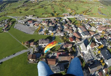 An aerial shot of a city with green meadows and residential houses. In the background, mountains and a colorful paraglider can be seen.