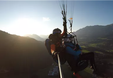 A person is gliding with a paraglider over a picturesque landscape. The sun is shining and the mountains are visible in the background.
