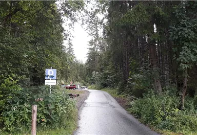 A narrow path leads through a forest with tall trees. On the left, a sign is visible, and in the background, some vehicles are parked.