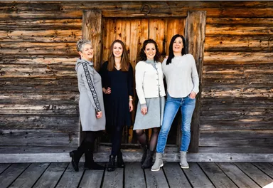Vier Frauen stehen fröhlich vor einer rustikalen Holzwand. Sie tragen verschiedene stylische Outfits und haben unterschiedliche Haarfrisuren.