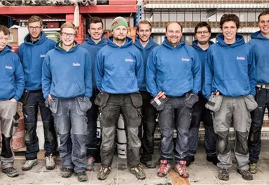 A group of ten craftsmen stands smiling in blue sweatshirts in front of shelves with work materials. They appear friendly and professional.