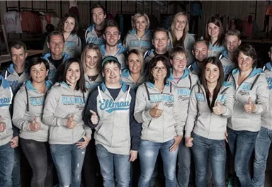 Eine Gruppe von lächelnden Menschen steht zusammen in einem Raum. Alle tragen Sweatshirts und zeigen den Daumen nach oben.