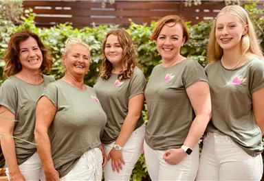 A group of five women is standing together and smiling in a friendly pose. They are wearing similar gray T-shirts and white pants, while plants are visible in the background.