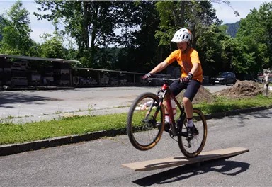 A cyclist jumps over a wooden board on a street. He is wearing a helmet and an orange shirt.