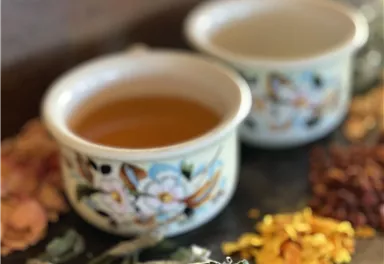 Zwei Tassen mit hellem Tee stehen auf einem Steinbrett. Um sie herum liegen verschiedene getrocknete Kräuter und Blüten.