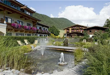Ein idyllischer Garten mit einem Teich und Sprudel. Im Hintergrund stehen zwei komfortable Gebäude, umgeben von Bergen und grüner Natur.