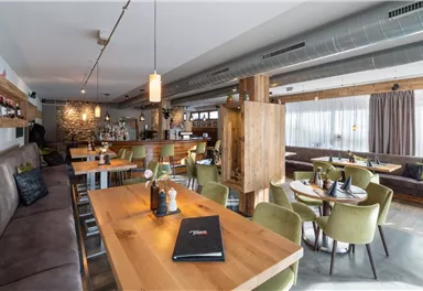 Ein modernes Restaurant mit Holztischen und gemütlichen, grünen Stühlen. Der Raum ist hell und einladend, mit einer Bar im Hintergrund.