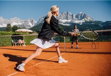 Eine Frau spielt Tennis auf einem Platz mit Blick auf beeindruckende Berge. Es ist sonnig und die Atmosphäre ist aktiv und sportlich.