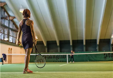 Eine Tennisspielerin steht auf dem Platz und blickt zum Netz. Der Innenbereich ist gut beleuchtet mit einer grünen Spielfläche.
