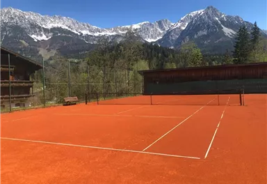 A tennis court with a red surface, surrounded by majestic mountains and a clear blue sky. Nature adds a picturesque backdrop.