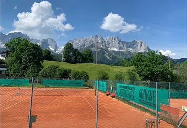 Zwei Tennisplätze mit rotem Sand und grünen Umzäunungen. Im Hintergrund erstrecken sich majestätische Berge unter einem klaren blauen Himmel.