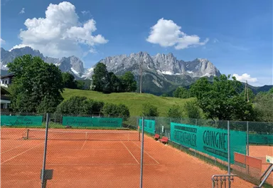 A tennis court surrounded by green trees and a picturesque mountain backdrop. The sky is blue with a few clouds.