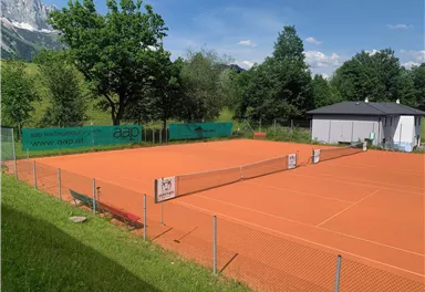 A tennis court made of sand with a clear blue sky in the background. Surrounded by trees and a small sports facility.