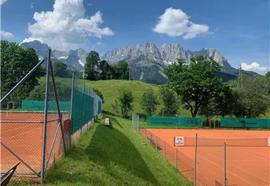 A tennis court surrounded by green meadows and trees. In the background, majestic mountains and a clear blue sky can be seen.