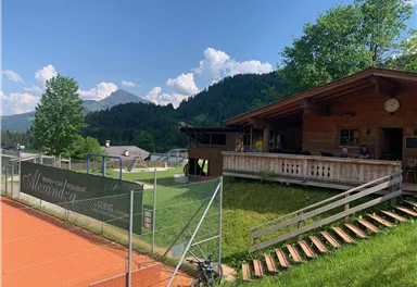 A wooden terrace overlooking a sports field and mountains in the background. It is sunny and the vegetation is green.