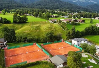 A tennis facility with several clay courts surrounded by green meadows and mountains in the background. The landscape is peaceful and inviting.