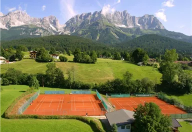 Eine wunderschöne Tennisanlage mit roten Plätzen inmitten einer grünen Landschaft. Im Hintergrund erheben sich majestätische Berge unter einem blauen Himmel.