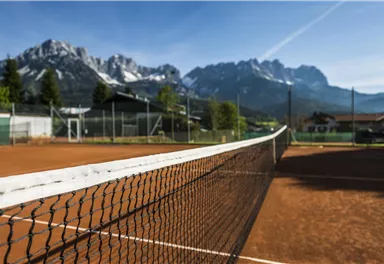 Ein Tennisplatz mit rotem Sand und einem klaren blauen Himmel. Im Hintergrund erheben sich beeindruckende Berge.