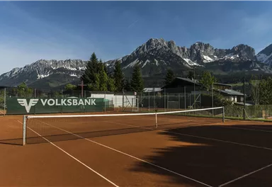 Ein Tennisplatz mit rotem Sand, umgeben von Bergen und grünen Bäumen. Im Hintergrund ist ein klarer Himmel zu sehen.