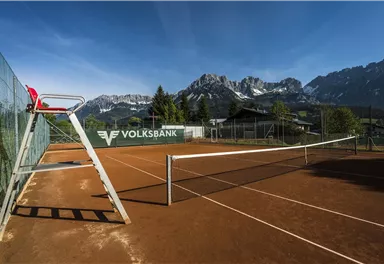 Ein Tennisplatz mit rotem Sand und einem Stuhl für den Schiedsrichter. Im Hintergrund sind beeindruckende Berge und ein blauer Himmel zu sehen.