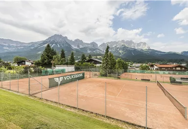 Ein Tennisplatz aus rotem Sand mit Blick auf majestätische Berge. Die Umgebung ist grün und bietet eine ruhige, alpine Atmosphäre.