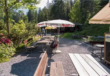 Ein ruhiger Außenbereich mit Holztischen und Bänken, umgeben von Bäumen. Ein Sonnenschirm bietet Schatten und lädt zum Verweilen ein.