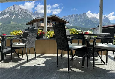 Eine gemütliche Terrasse mit schwarzen Stühlen und Tischen. Im Hintergrund sind majestätische Berge und ein blauer Himmel zu sehen.