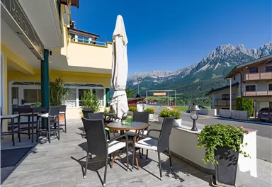 Ein gemütlicher Außenbereich mit Tischen und Stühlen. Im Hintergrund sind schöne Berge und ein strahlend blauer Himmel zu sehen.