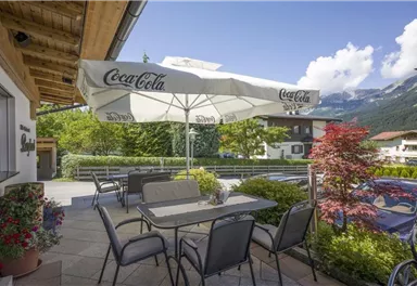 Eine gemütliche Terrasse mit einem großen Sonnenschirm und Sitzplätzen. Im Hintergrund sind grüne Bäume und Berge zu sehen.