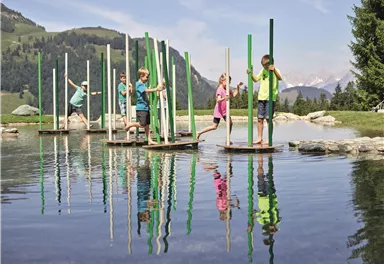 Fünf Kinder stehen auf Plattformen im Wasser und balancieren mit bunten Stangen. Im Hintergrund sind grüne Hügel und ein klarer Himmel zu sehen.