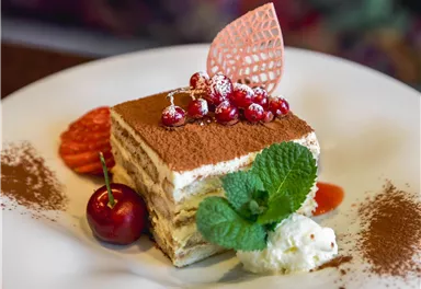 Ein köstliches Stück Tiramisu auf einem weißen Teller, garniert mit roten Früchten und frischen Minzblättern. Im Hintergrund sieht man eine bunte, unscharfe Textur.