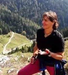 Eine Person steht auf einem Berg mit grünen Wäldern im Hintergrund. Der Himmel ist teilweise bewölkt und die Landschaft ist hügelig.